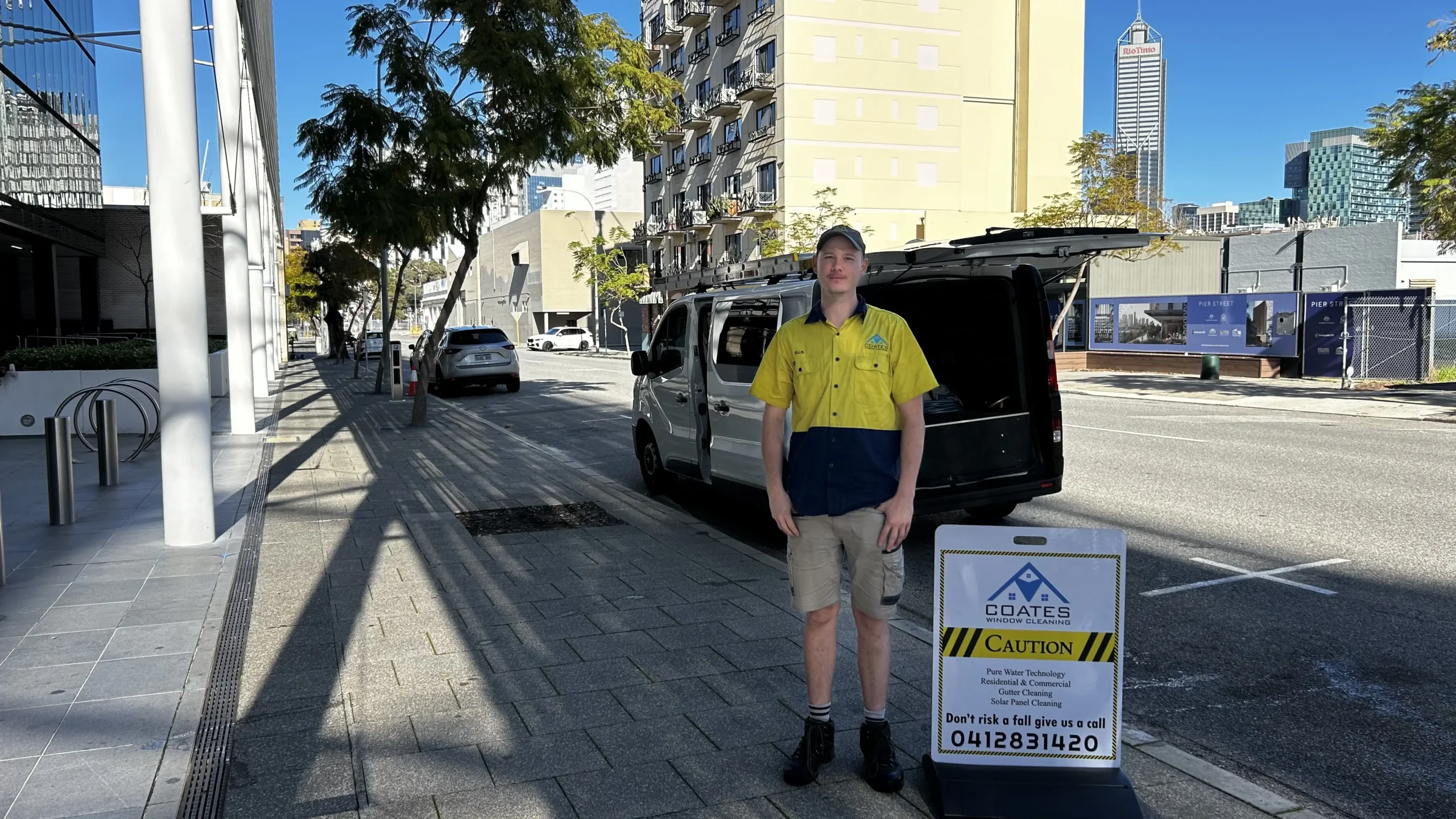 A person in a yellow and navy work uniform stands on a sidewalk next to a parked van, beside a caution sign from a company named "Coates" with a contact number displayed. Urban buildings are visible.