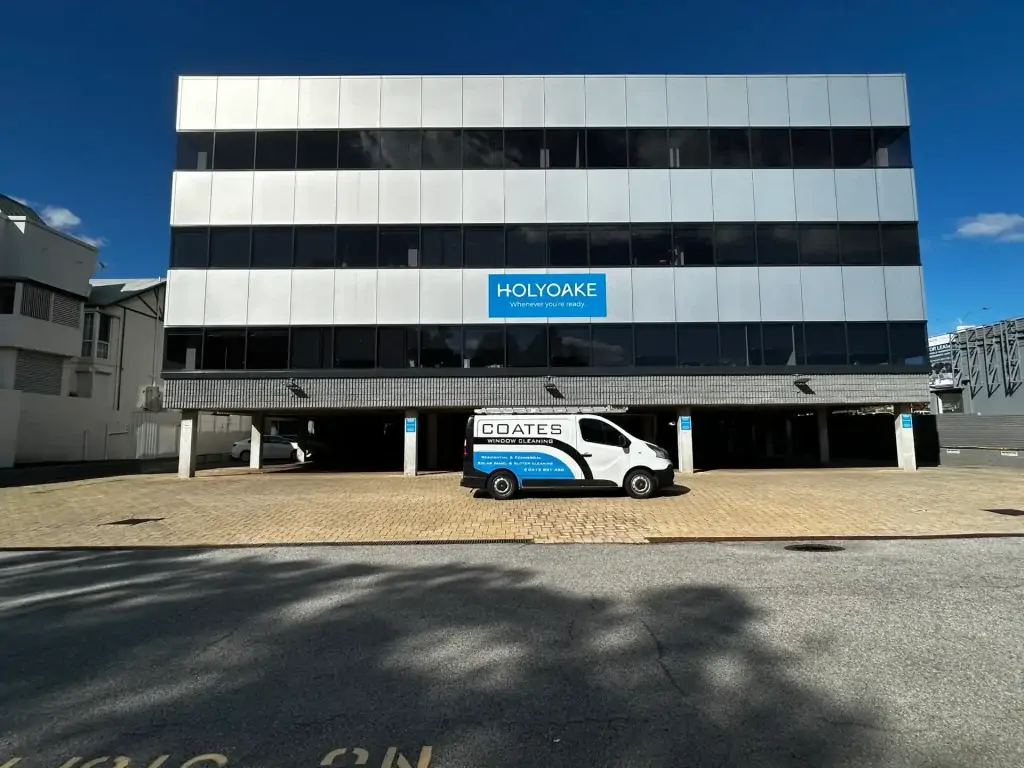 A white and gray office building with a "Holyoake" sign on the facade. A Coates service van, possibly for window cleaning, is parked in front on a brick driveway under a blue sky.