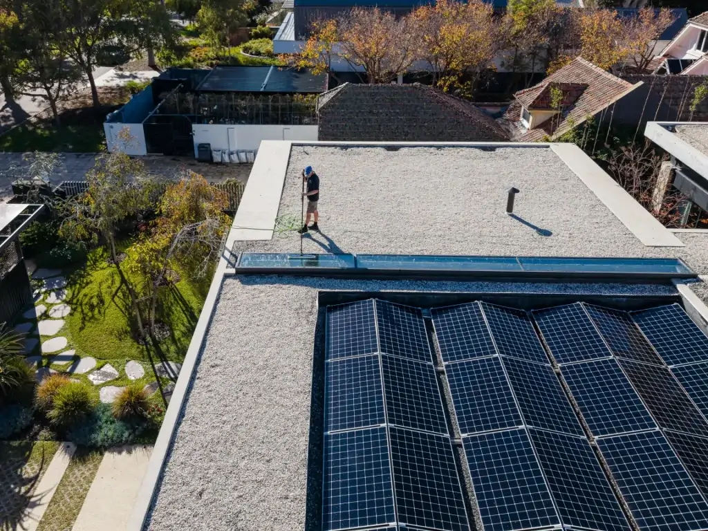 A person uses a hose to clean a flat rooftop with solar panels, surrounded by trees and nearby buildings on a sunny day—a scene reminiscent of expert window cleaning in Perth.