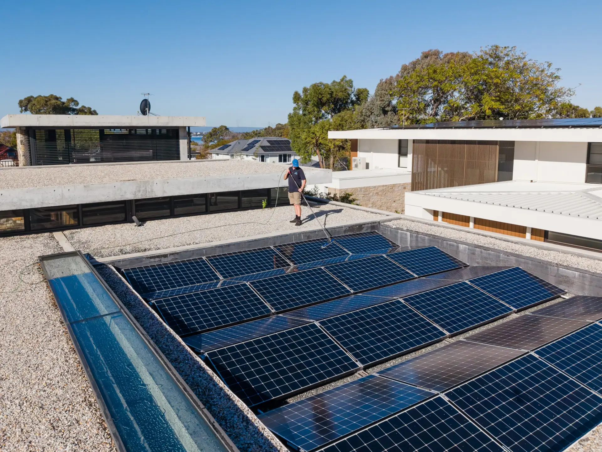 A person stands on a rooftop beside an array of solar panels on a sunny day, surrounded by modern buildings and trees, showcasing the pristine results often achieved by professional window cleaning Perth services.