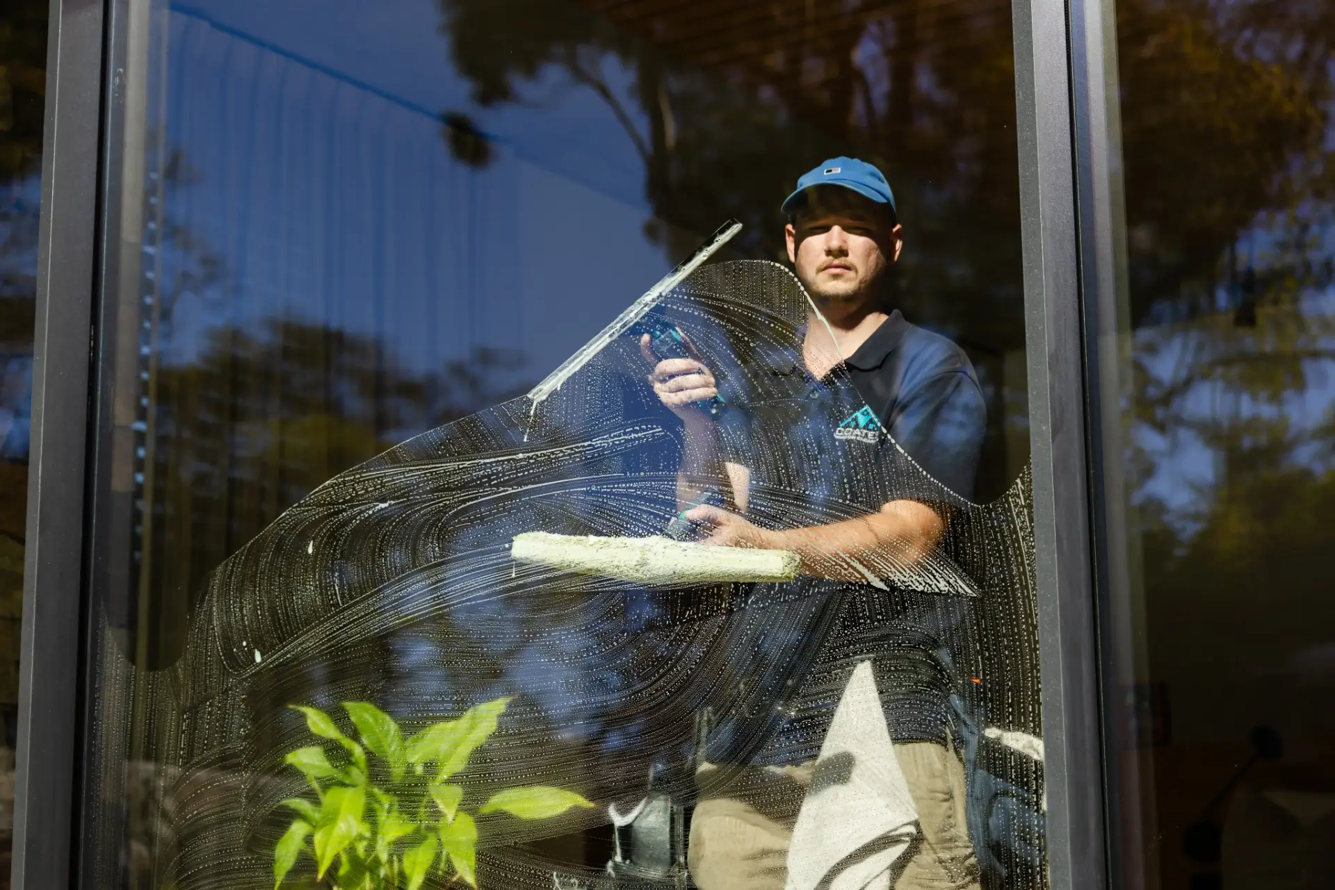 A person wearing a blue cap cleans a large glass window with a squeegee and cleaning tool, demonstrating professional Window Cleaning Perth standards. A green plant is visible inside near the window, while outdoor reflections appear on the glass.
