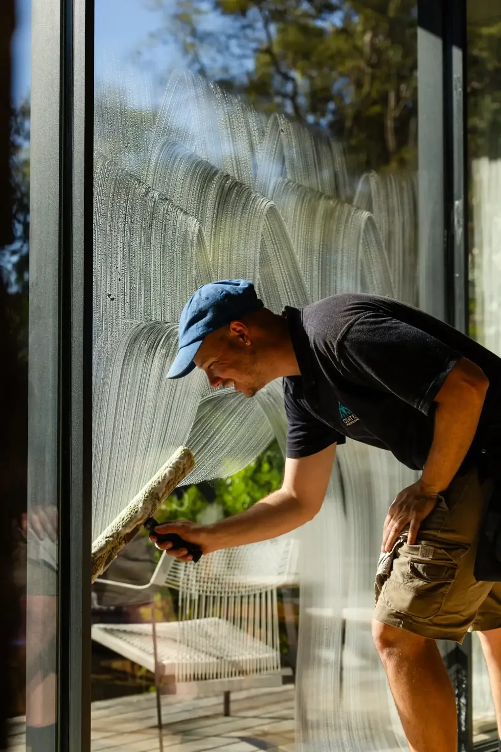 A man wearing a blue cap and dark shirt cleans a large glass window with a squeegee, providing professional Window Cleaning Perth services. Outdoor furniture and trees are visible through the sparkling glass.