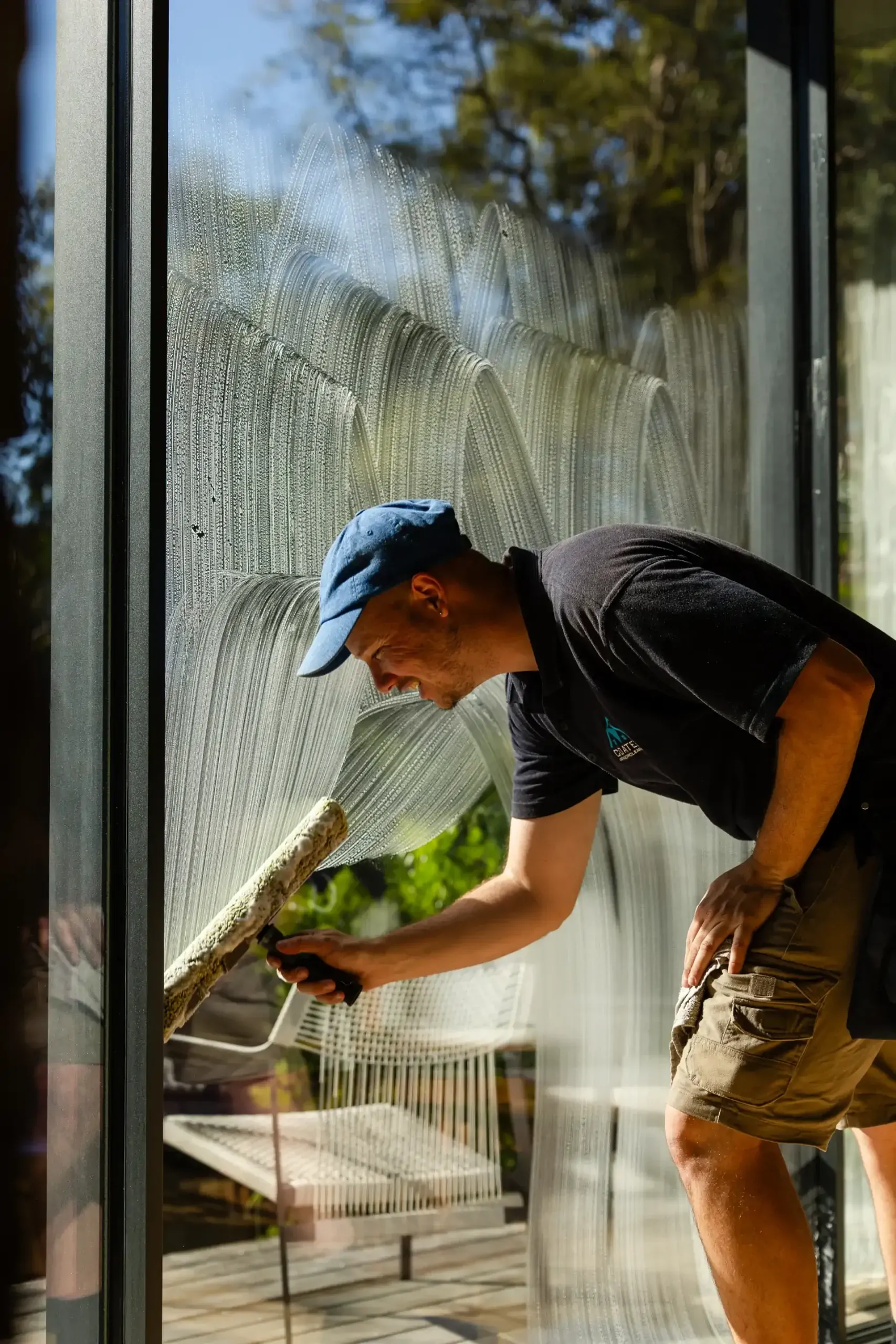 A man wearing a blue cap and dark shirt cleans a large glass window with a squeegee, providing professional Window Cleaning Perth services. Outdoor furniture and trees are visible through the sparkling glass.