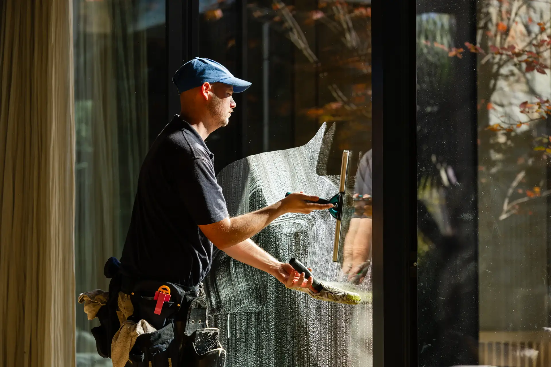 A man wearing a blue cap and tool belt is cleaning a large glass window with a squeegee on a sunny day, providing expert Window Cleaning Perth services.