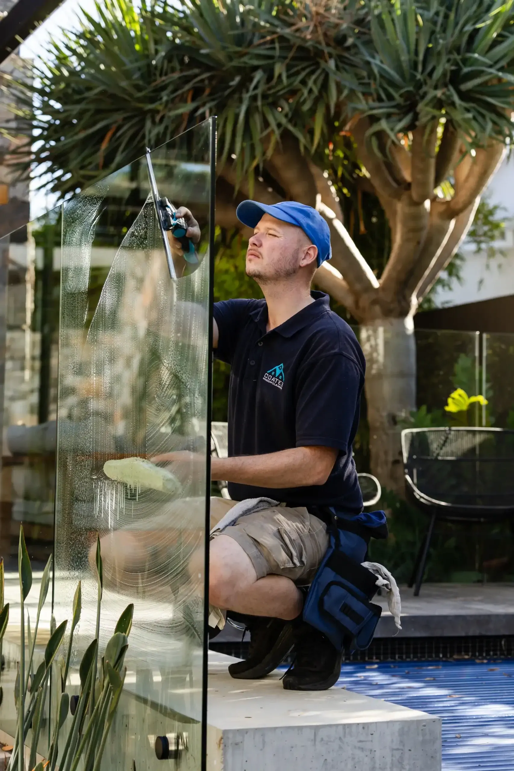 A man in a blue cap and black polo shirt kneels and cleans a glass panel outdoors with a squeegee, demonstrating professional window cleaning Perth standards near a modern pool area with plants and patio furniture in the background.