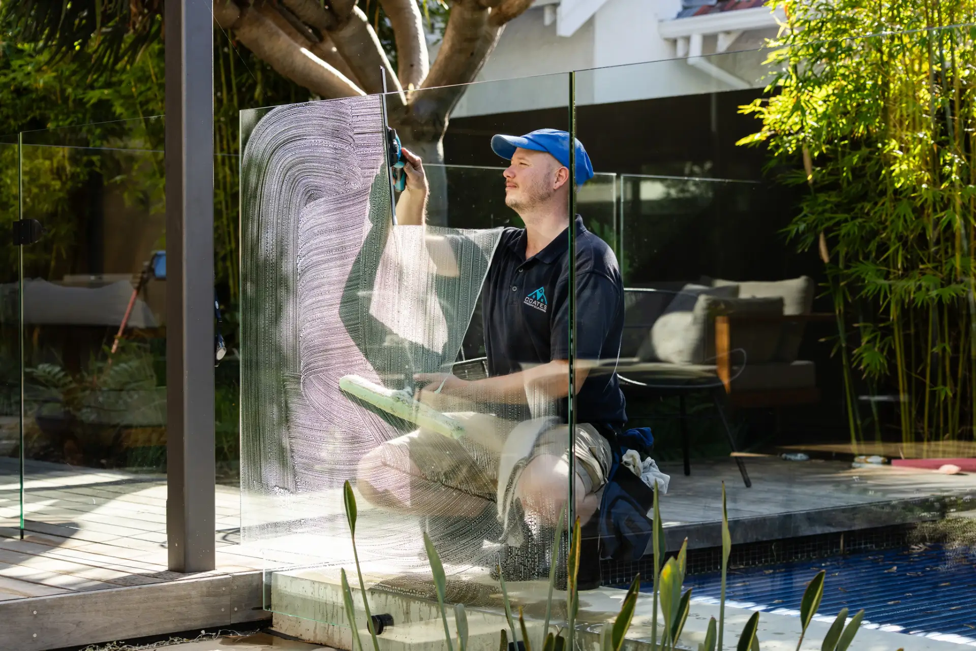 A man wearing a blue cap and black shirt kneels by a pool while cleaning a glass fence with a squeegee in a backyard setting, showcasing the attention to detail often seen in Window Cleaning Perth services.
