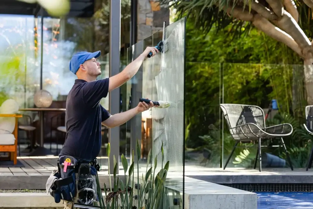 A man wearing a blue cap and tool belt cleans an outdoor glass fence with a squeegee near a modern house and garden, showcasing professional window cleaning Perth services.