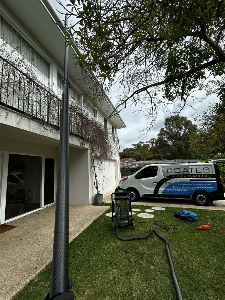 A window cleaning pole is extended to a second-story window of a white house, with a Coates Window Cleaning van and equipment—specializing in gutter cleaning Perth—on the grass nearby.