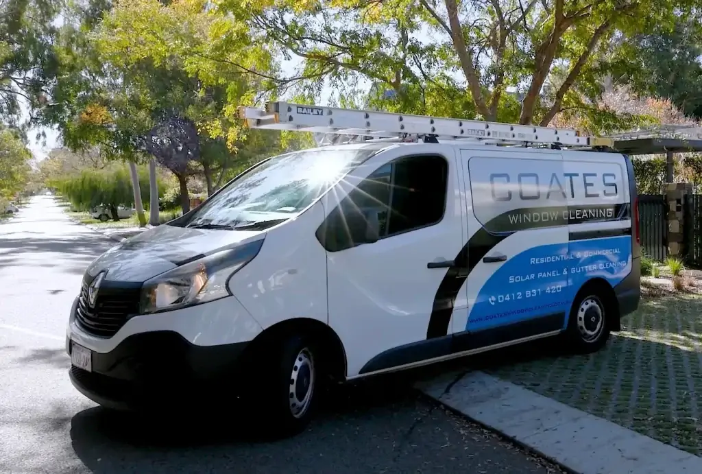 A white van with a ladder on top is parked on a street, proudly displaying the Coates Window Cleaning logo and contact details on its side. Trees and sunlight are visible in the background.