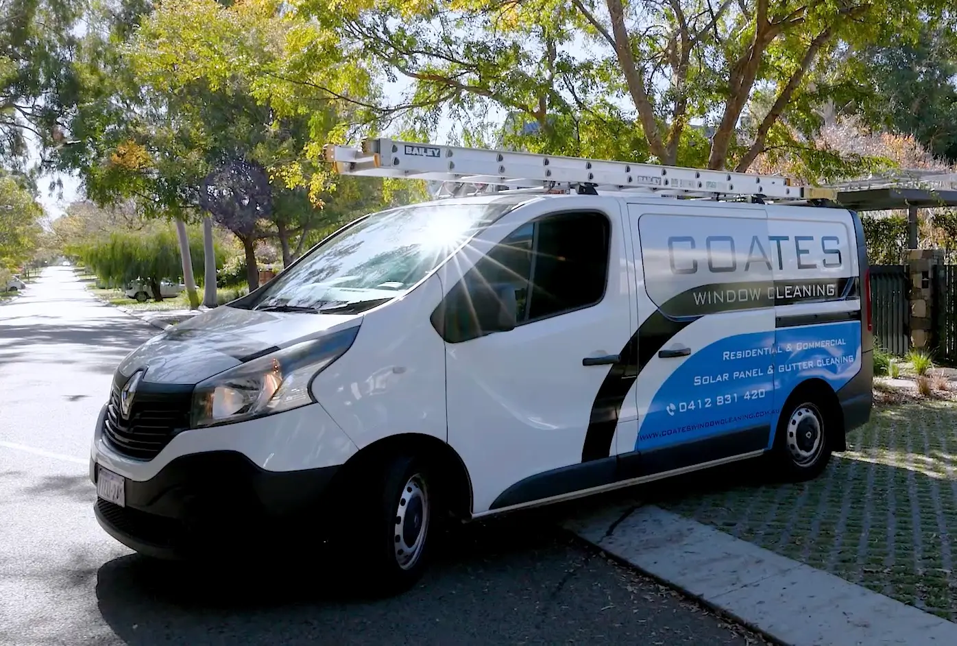 A white van with a ladder on top is parked on a street, proudly displaying the Coates Window Cleaning logo and contact details on its side. Trees and sunlight are visible in the background.